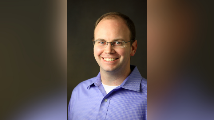 Man with glasses and short hair wearing a light purple collared shirt, smiling in front of a dark background.