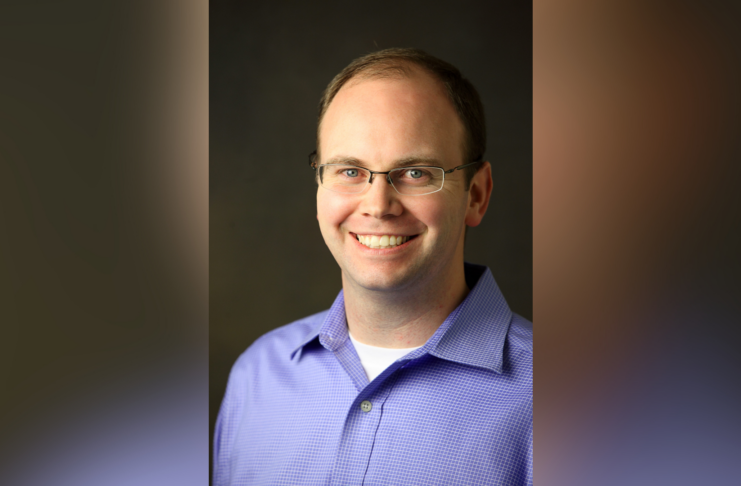 Man with glasses and short hair wearing a light purple collared shirt, smiling in front of a dark background.