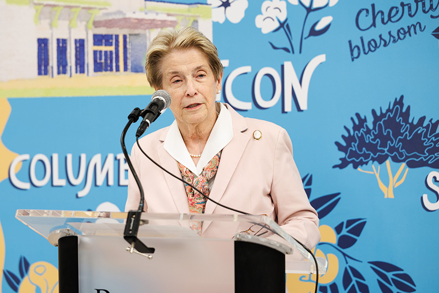 A woman in a light suit speaks at a podium in front of a colorful, decorated backdrop.