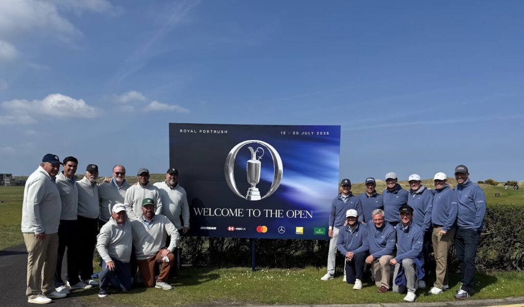 A group of people pose in front of a Welcome to The Open sign at a golf course on a sunny day.