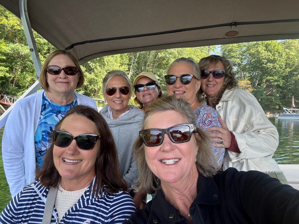 Seven women wearing sunglasses smile for a group selfie on a boat with trees and water in the background.