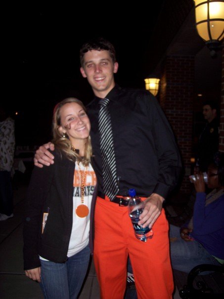Two people pose together outdoors at night; the man wears orange pants and holds a water bottle.