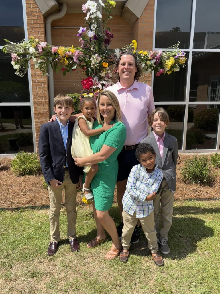A family of six poses outside in front of a flower-decorated cross, dressed in spring attire.