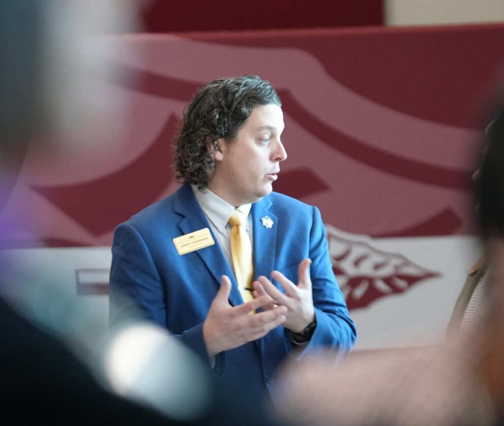 A man in a blue suit and yellow tie speaks, gesturing with his hands, in front of a maroon and white wall.