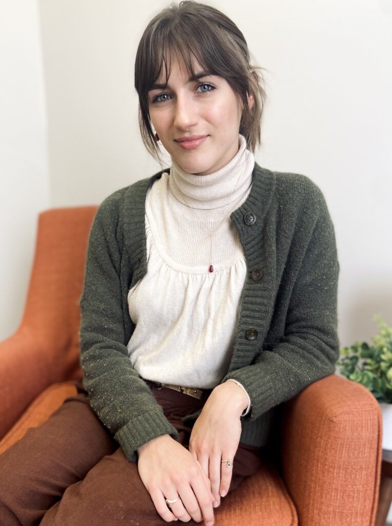 Woman with short brown hair sits on an orange chair, wearing a green cardigan, white top, and brown pants.