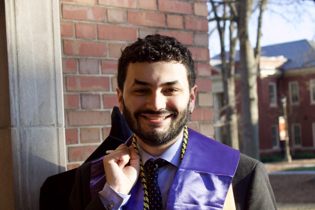 A man wearing graduation attire stands outside near a brick building and smiles at the camera.