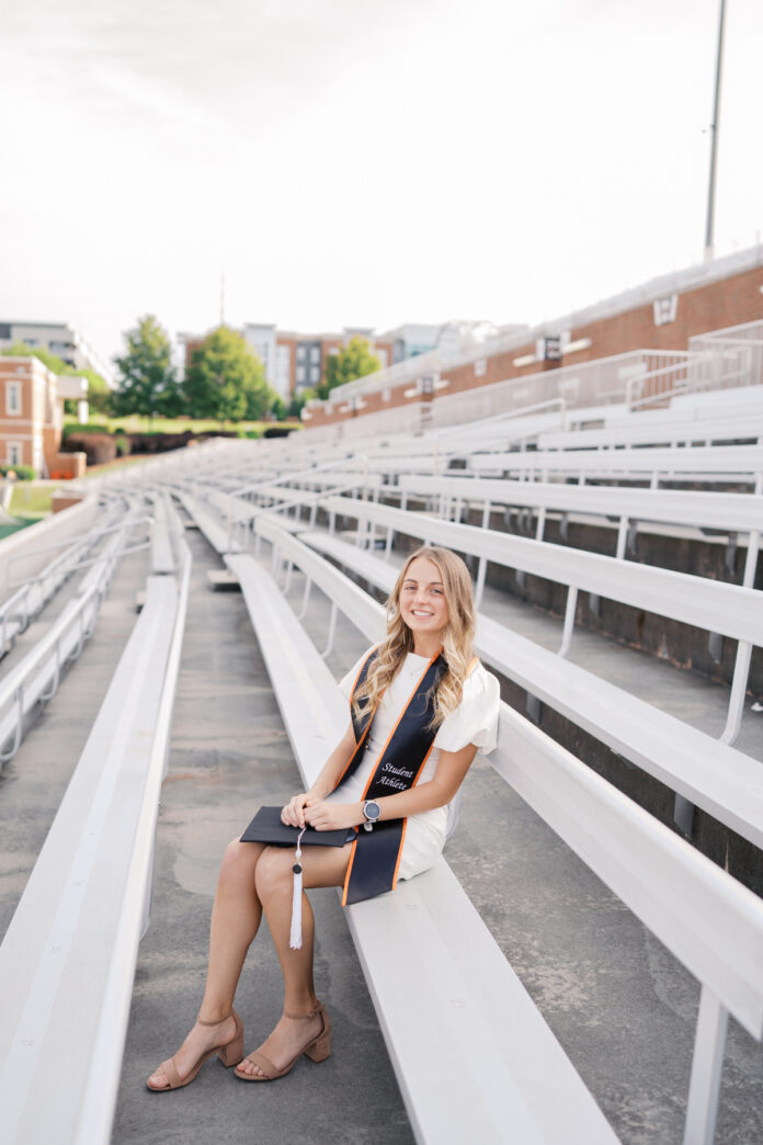 Reese Robertson, in a graduation stole and dress, sits on stadium bleachers, holding her cap and smiling at the camera.