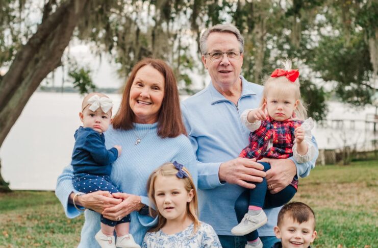 Two adults and four young children pose for a family photo outdoors on grass with trees and water in the background.