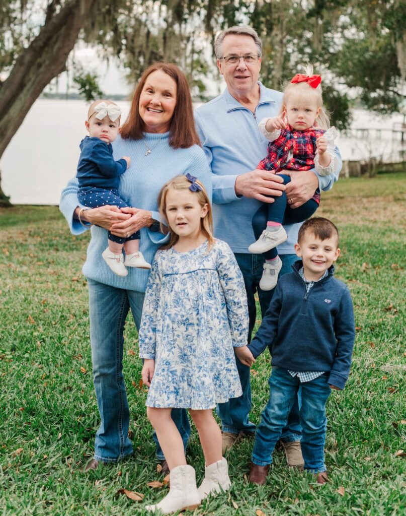 Two adults and four young children pose for a family photo outdoors on grass with trees and water in the background.