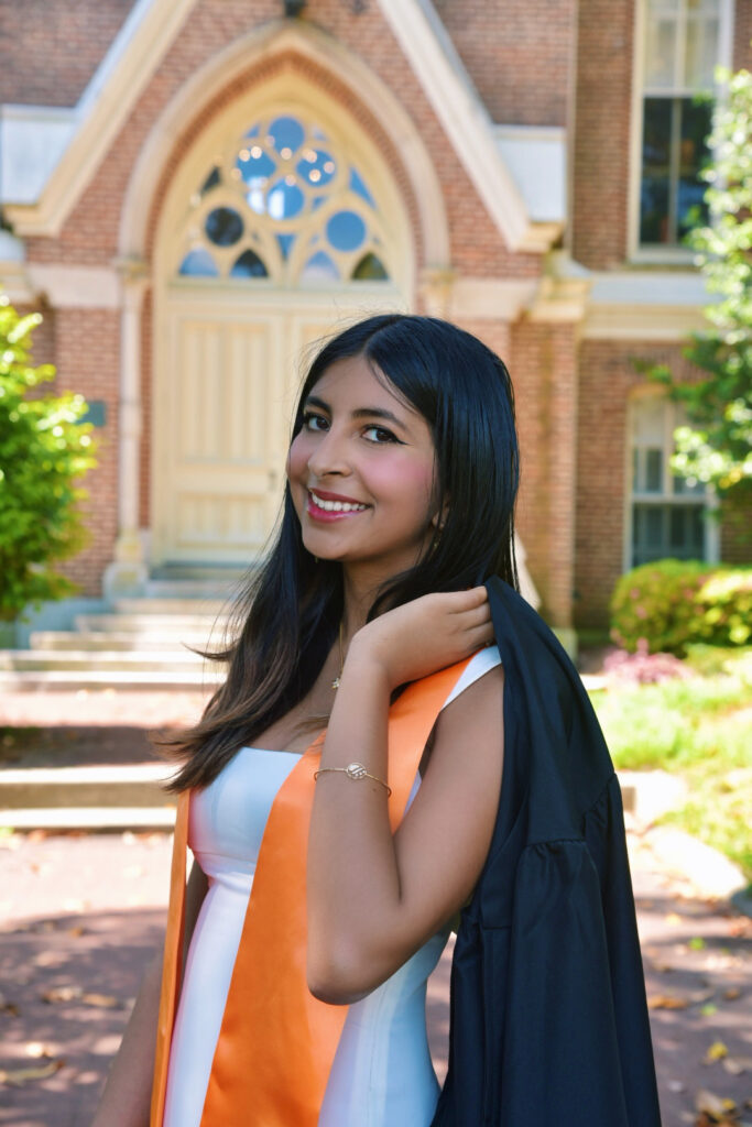 Tanvi Sarma in graduation attire stands outdoors, holding her gown over her shoulder in front of a brick building.