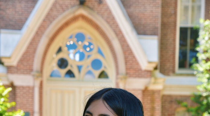 Graduate Spotlight: Tanvi Sarma Tanvi Sarma in graduation attire stands outdoors, holding her gown over her shoulder in front of a brick building.