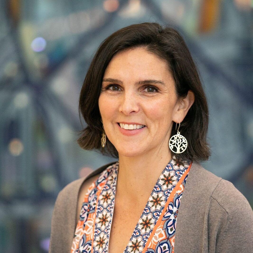 A woman with short dark hair, wearing a patterned blouse, cardigan, and tree-shaped earrings, smiles at the camera.