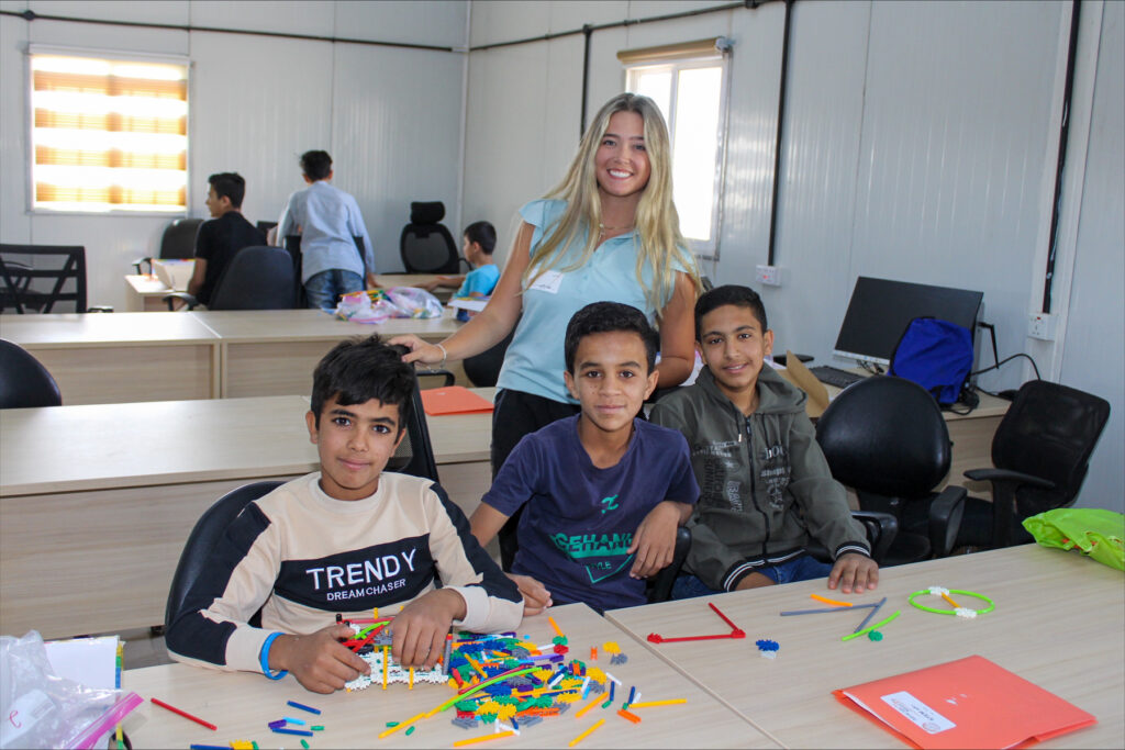 Three boys sit at a table with building toys while Halley Smith stands behind them, all smiling in a classroom setting.