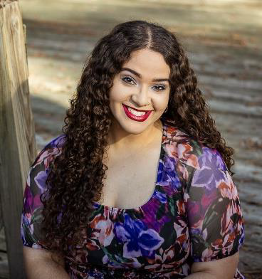 Woman with long curly hair and floral dress smiling outdoors, seated near a wooden post with sunlight in the background.
