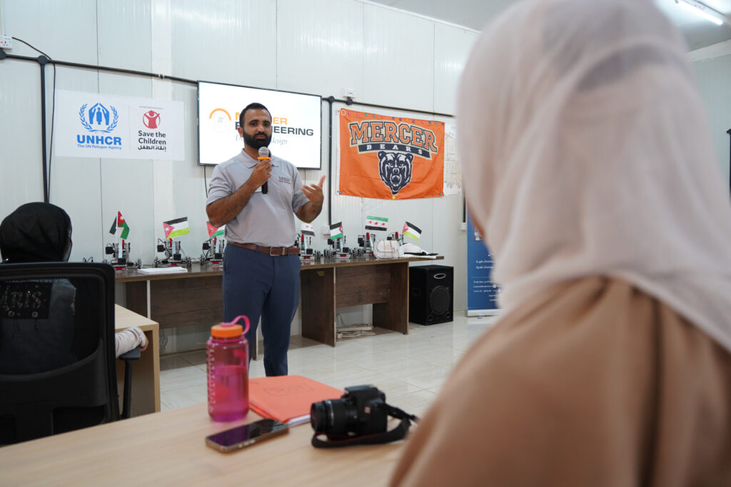 A man speaks into a microphone in a classroom with UNHCR and Mercer banners, addressing seated attendees.