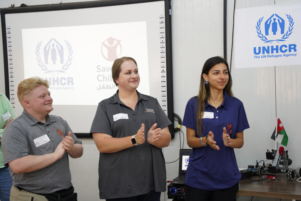 Three people stand and clap at an indoor event with UNHCR and Save the Children logos displayed in the background.