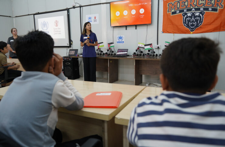 A woman presents to a classroom of students with UNHCR and Save the Children logos displayed on the screen.