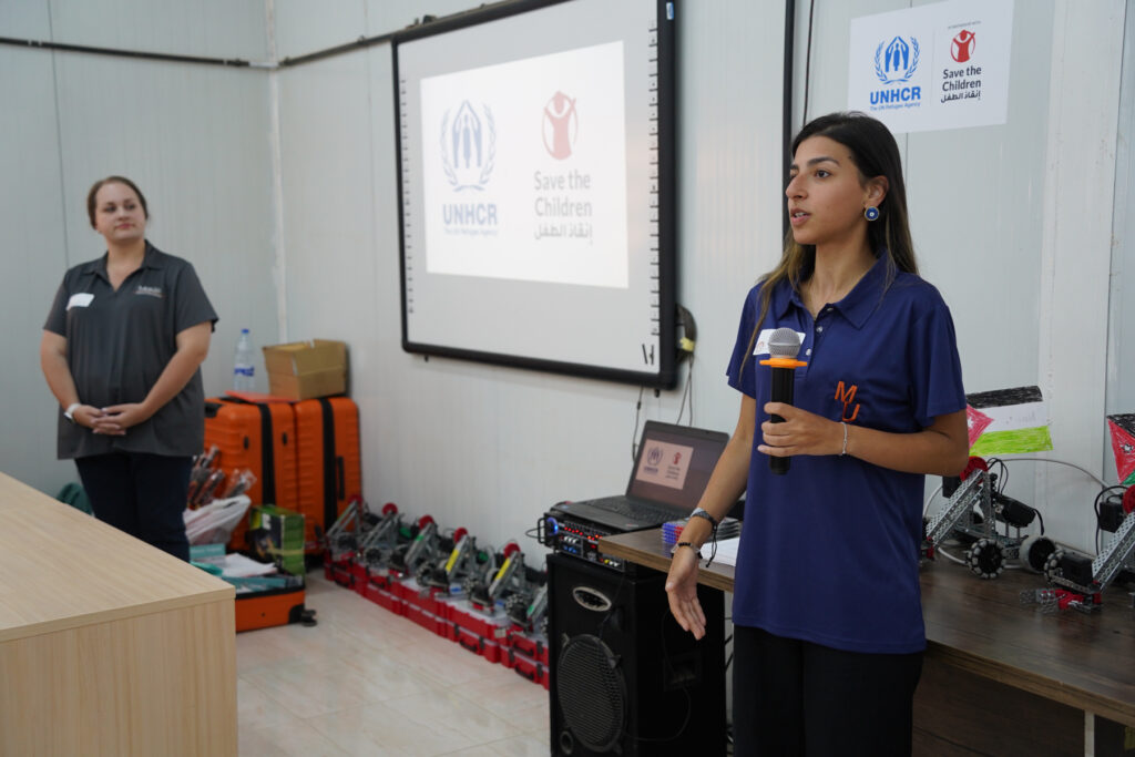Two women stand in a classroom, one speaking, with UNHCR and Save the Children logos displayed on a screen behind them.