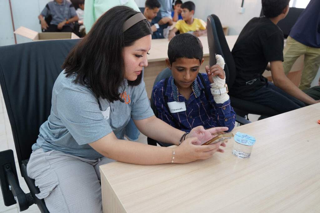 A woman shows a boy with a bandaged arm something on her phone while sitting at a table in a classroom.