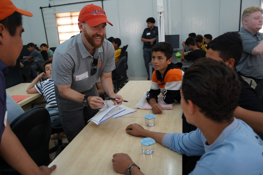 Man in a grey shirt and orange cap interacts with seated boys at a classroom table, others observe in the background.
