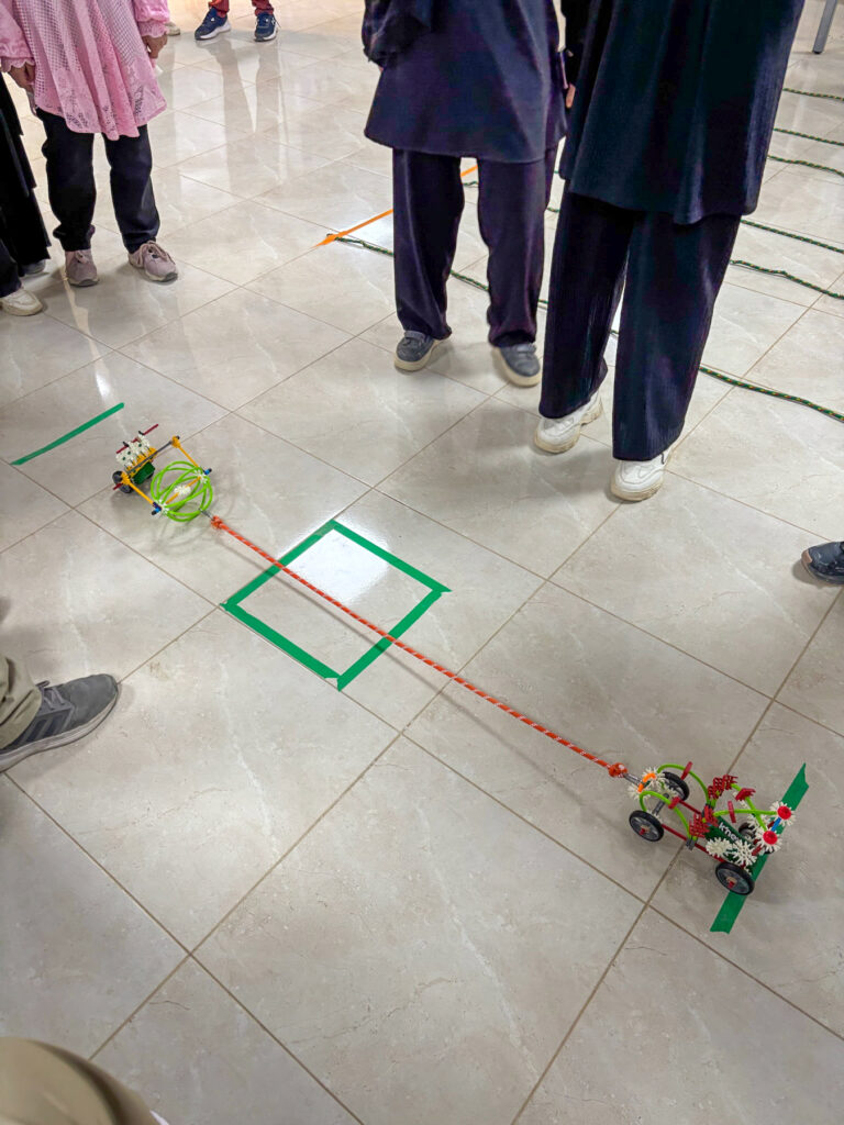Two small wheeled robots, connected by a red string, are on a tiled floor surrounded by people standing nearby.