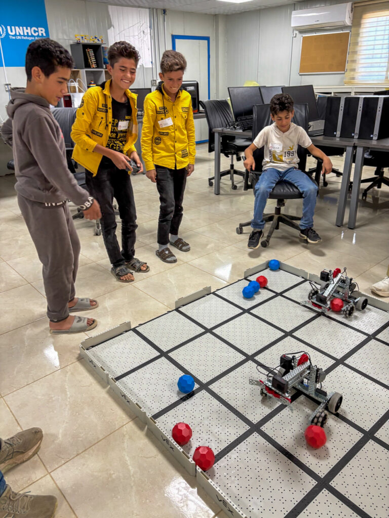 Four boys watch and control small robots on a grid while another boy is seated in a room with computers and a UNHCR sign.