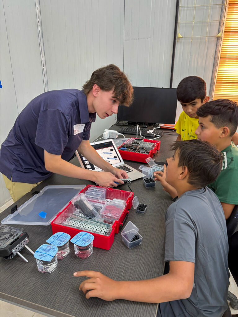 A young instructor shows three boys how to use robotics kits at a table with containers, tools, and a computer.