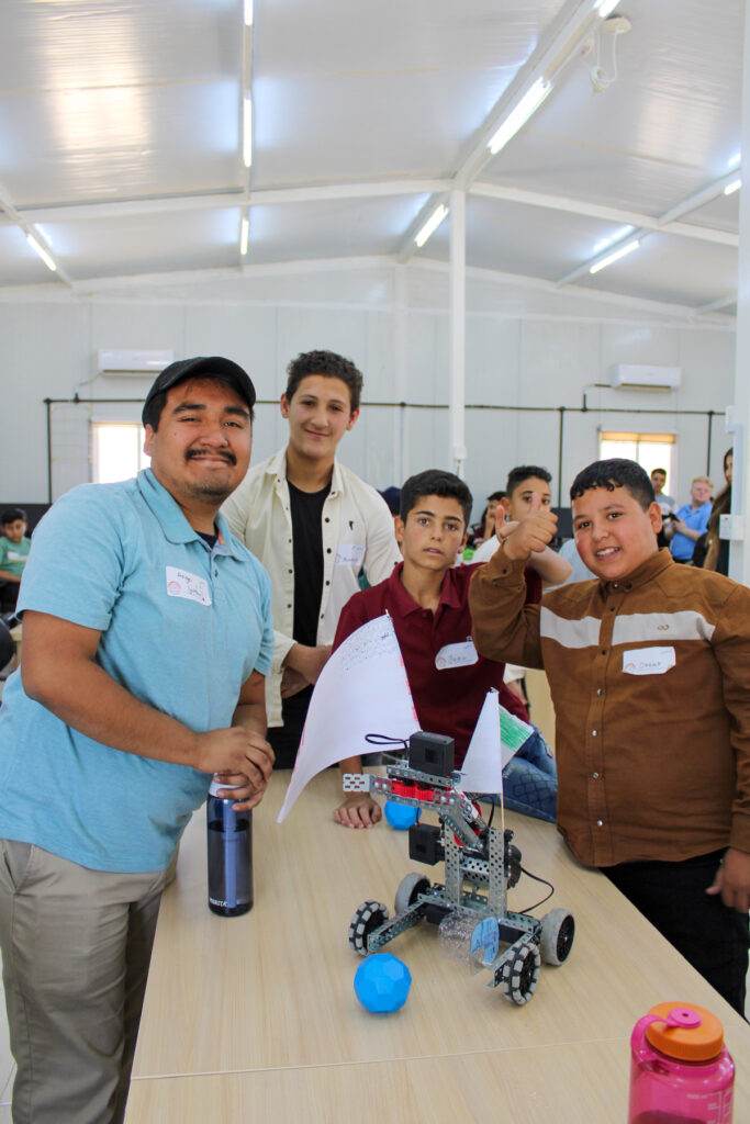 Four boys stand around a table with a small robot, smiling and giving a thumbs-up in a classroom setting.