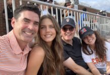 Love of golf leads to professional partnership for two alumni Four people sit closely together in a stadium, smiling at the camera, with bleachers and other attendees in the background.