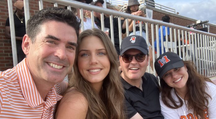 Love of golf leads to professional partnership for two alumni Four people sit closely together in a stadium, smiling at the camera, with bleachers and other attendees in the background.