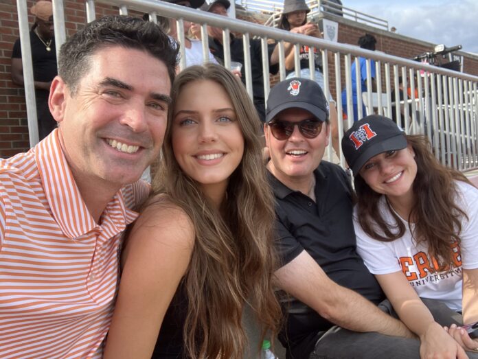 robert,_tom_and_girlfriend Four people sit closely together in a stadium, smiling at the camera, with bleachers and other attendees in the background.