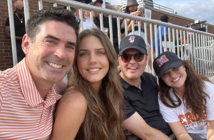 Four people sit closely together in a stadium, smiling at the camera, with bleachers and other attendees in the background.
