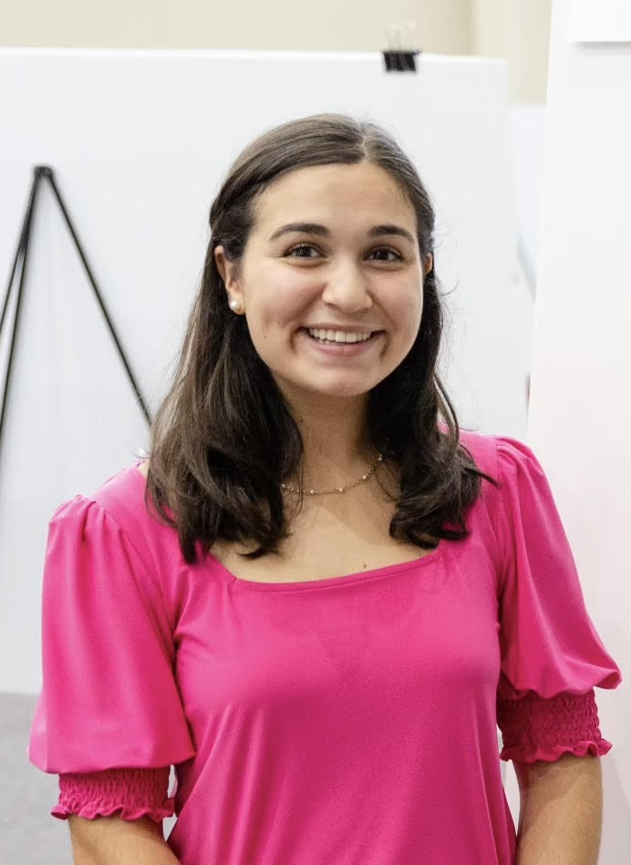 A woman with long dark hair smiles, wearing a bright pink top, standing indoors with a white background.
