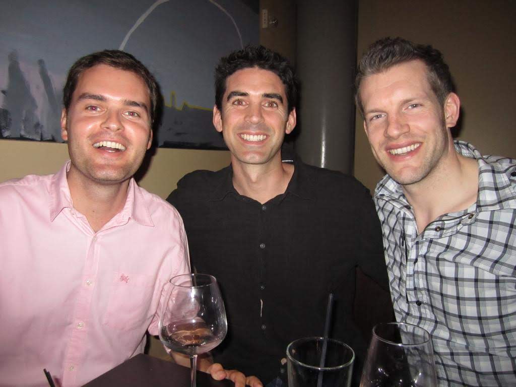 Three men sit at a table with empty wine glasses, smiling at the camera in a dimly lit indoor setting.