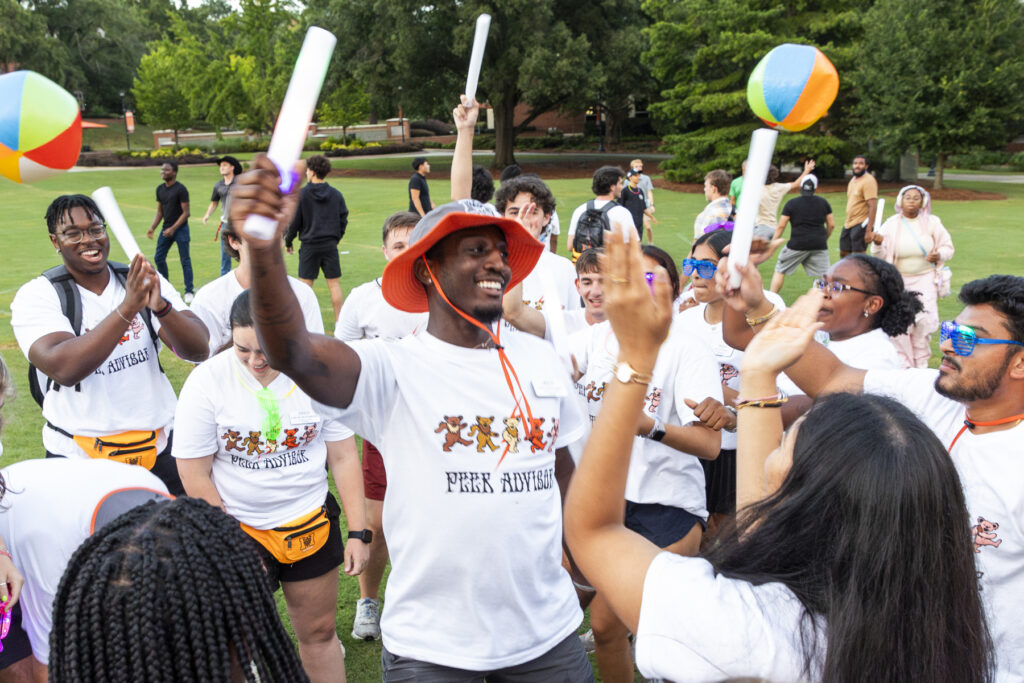 A group of people in matching shirts celebrate outdoors, holding papers and beach balls, surrounded by greenery.