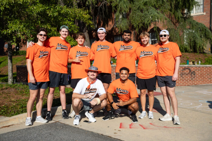 Eight people in matching orange Mercer shirts and one in white pose together outside on a sunny day.