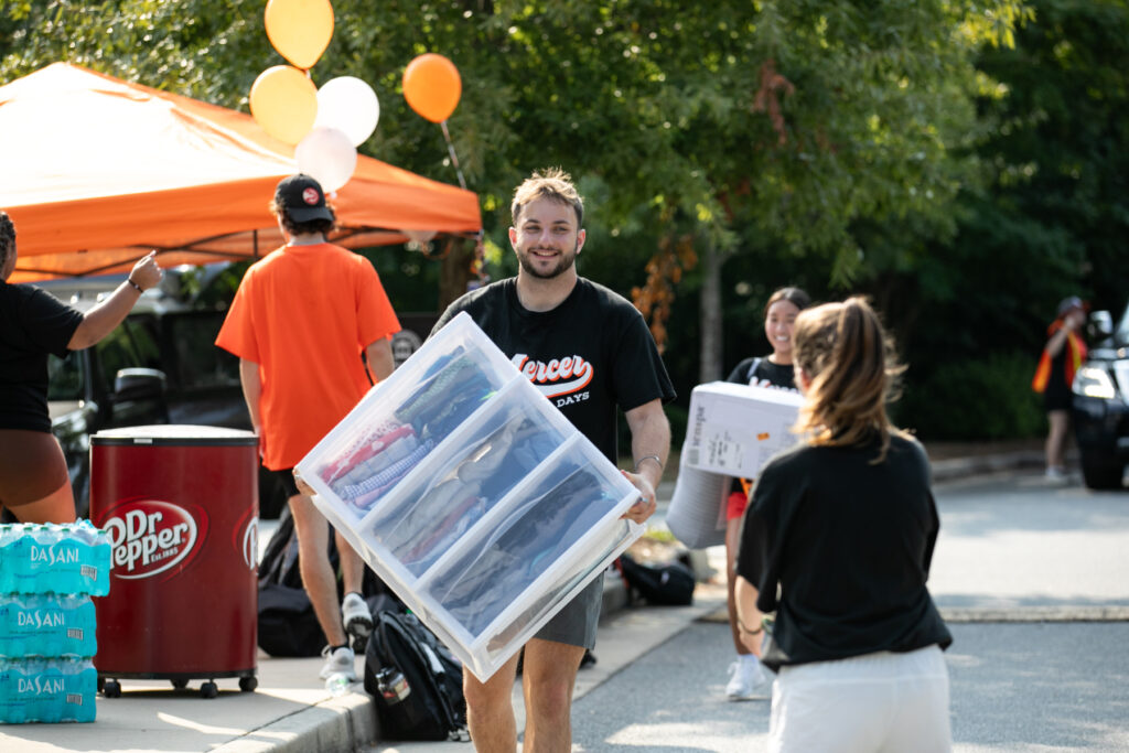 A man carries a large plastic storage bin outdoors while others move belongings during Mercer move-in.