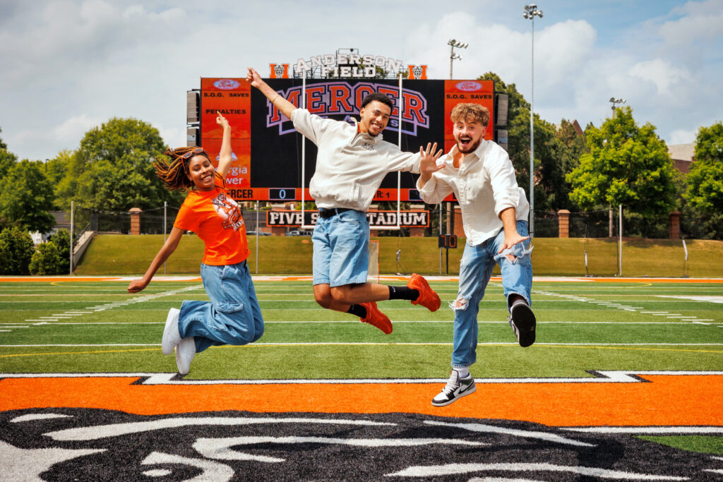 Three Mercer students jump in the air on a football field in front of a scoreboard.