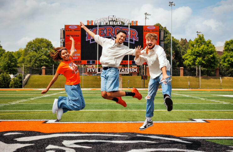 Three Mercer students jump in the air on a football field in front of a scoreboard.