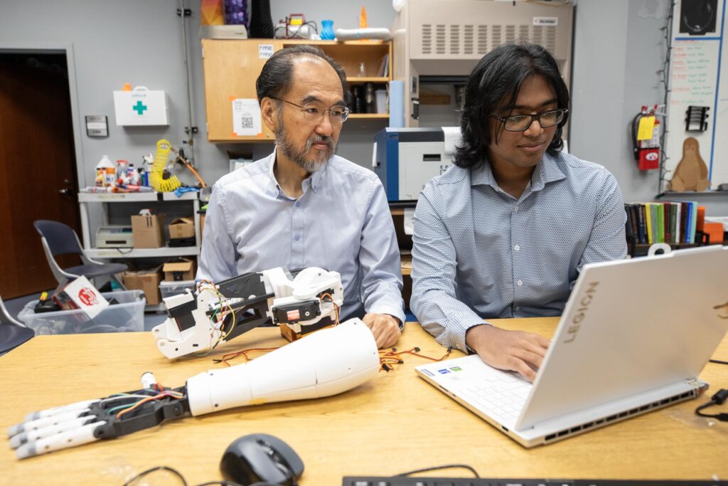 Two people sit at a desk with a laptop and robotic prosthetic hands in a laboratory setting.