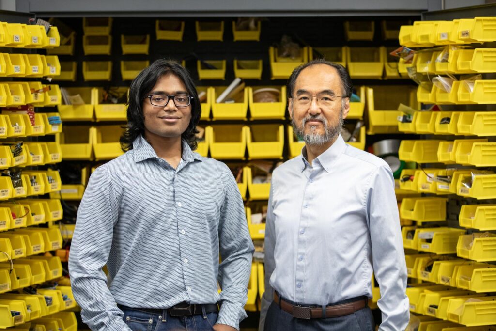 Two men standing in front of shelves with yellow storage bins filled with various items, both looking at the camera.