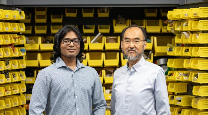 Summer research project explores alternative machine learning framework Two men standing in front of shelves with yellow storage bins filled with various items, both looking at the camera.
