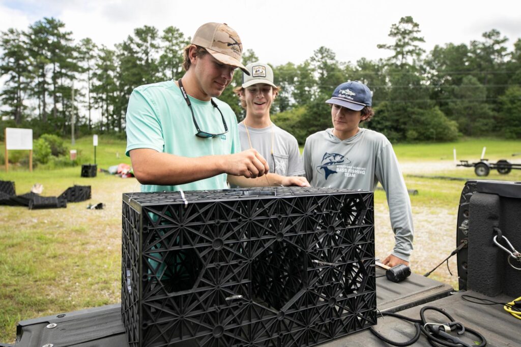 Three young men examine and assemble a black plastic crate outdoors, with trees and grass in the background.
