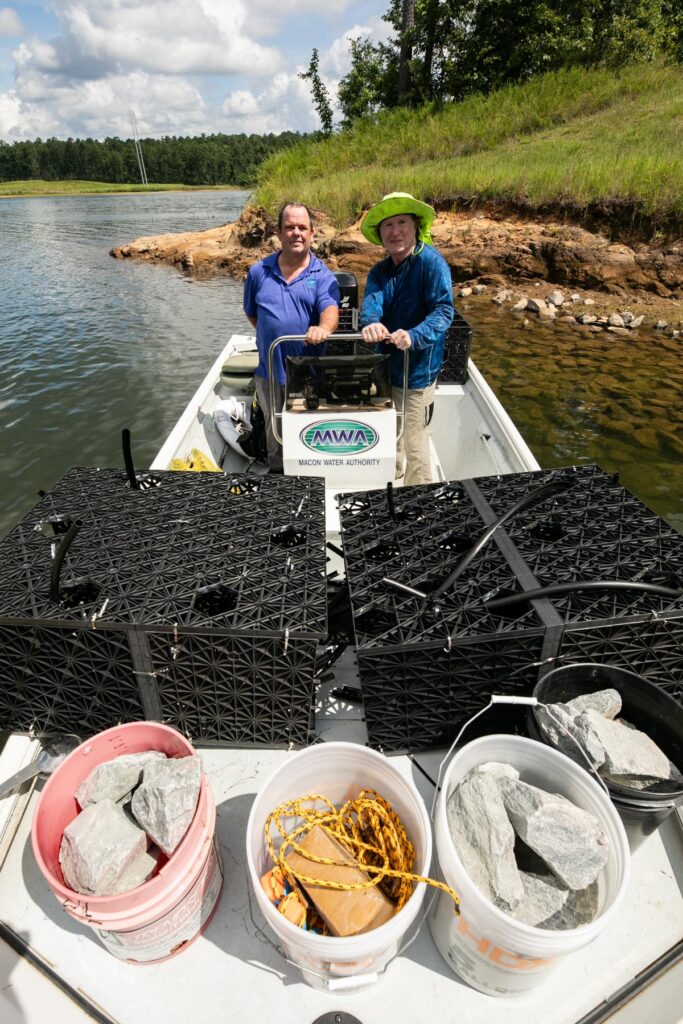 Two people on a boat with buckets of rocks and black crates near a grassy shoreline under a partly cloudy sky.