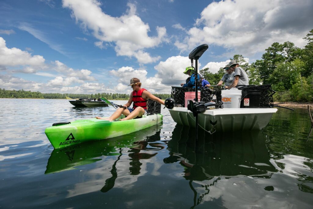 A person in a green kayak approaches a small motorboat with three people on a calm lake under a partly cloudy sky.