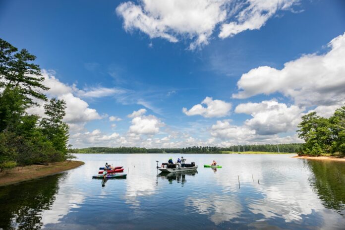 2507_MURS_MWA_CiSmith-342 Several people on small boats and kayaks are floating on a calm lake surrounded by trees under a partly cloudy sky.