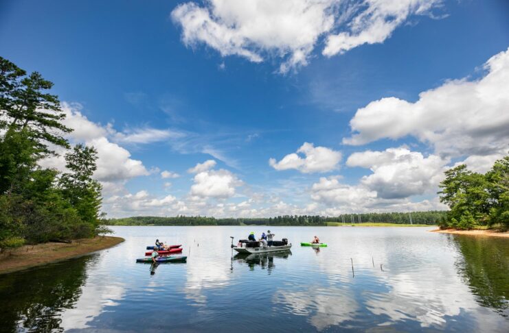 Several people on small boats and kayaks are floating on a calm lake surrounded by trees under a partly cloudy sky.