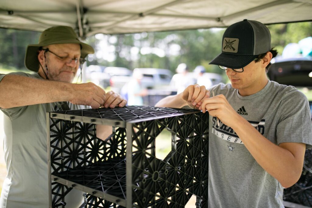 Two people assemble a black plastic crate outdoors under a canopy, focusing closely on connecting its pieces.