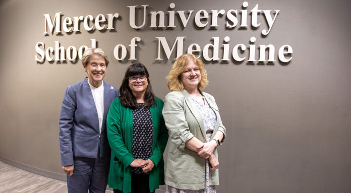 Dr-House-Den-Photo-JUL2025 Three women stand smiling in front of a wall sign that reads Mercer University School of Medicine.