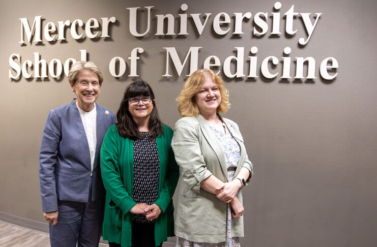 Three women stand smiling in front of a wall sign that reads Mercer University School of Medicine.